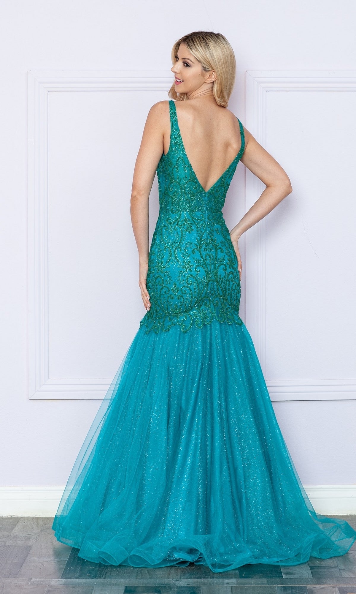 A woman stands against a light-colored wall wearing the Low V-Neck Long Glitter Mermaid Prom Dress 9388, a fitted sleeveless teal gown with sparkling embroidery and a deep V-back.